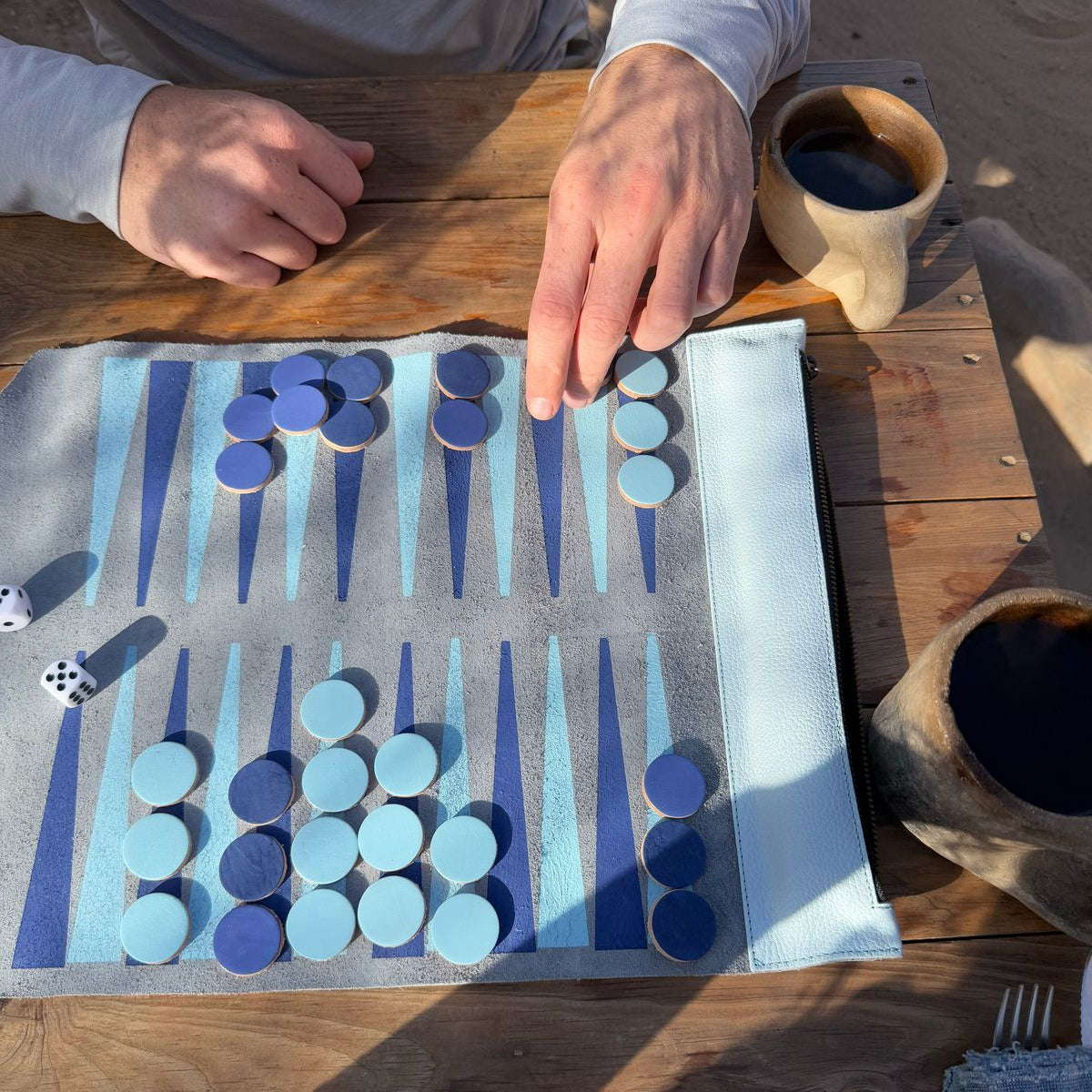 Backgammon game in progress on a wooden table with a person's hands and a cup of coffee.